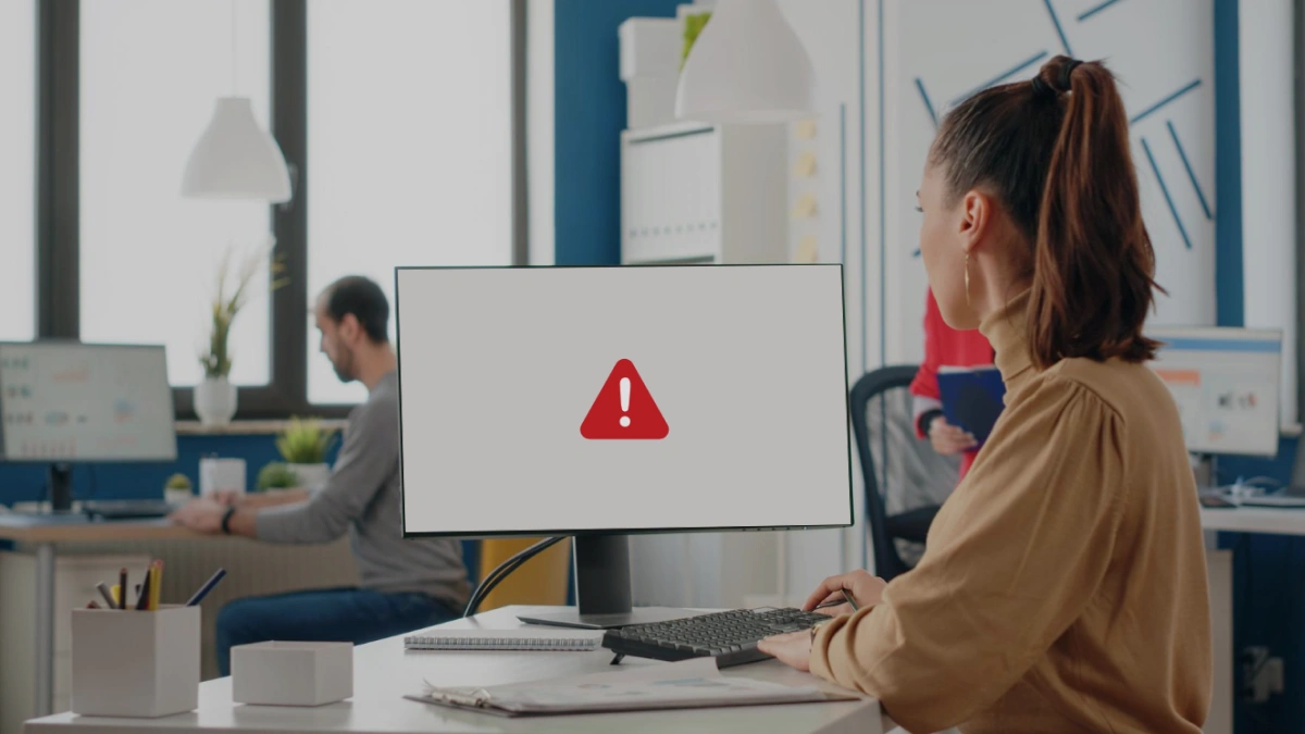 Woman in office setting looking at desktop monitor with error icon on screen.